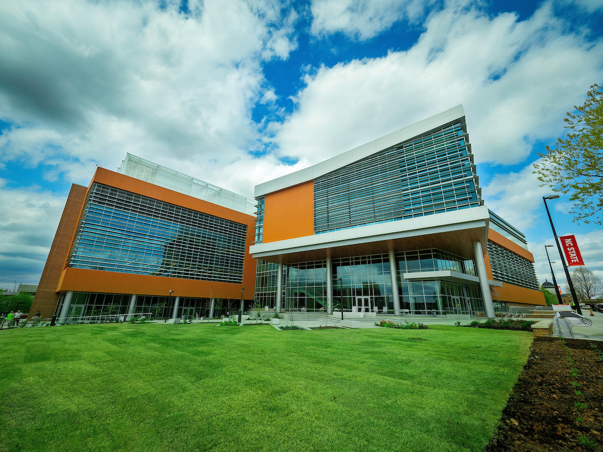 Modern brick and glass university building against backdrop of blue sky with white, fluffy clouds, and green grass.