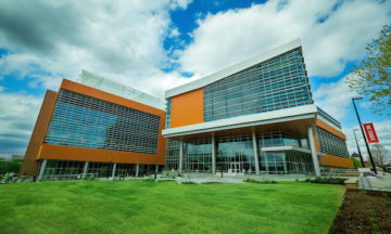 Modern brick and glass university building against backdrop of blue sky with white, fluffy clouds, and green grass.